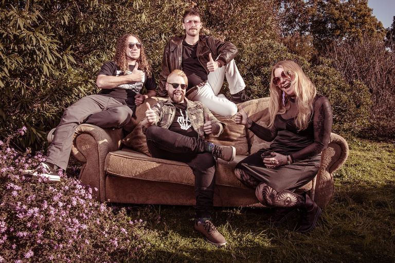 Members of blues rock band Kitchen Witch giving thumbs up while sitting on a couch in an outdoor garden setting.